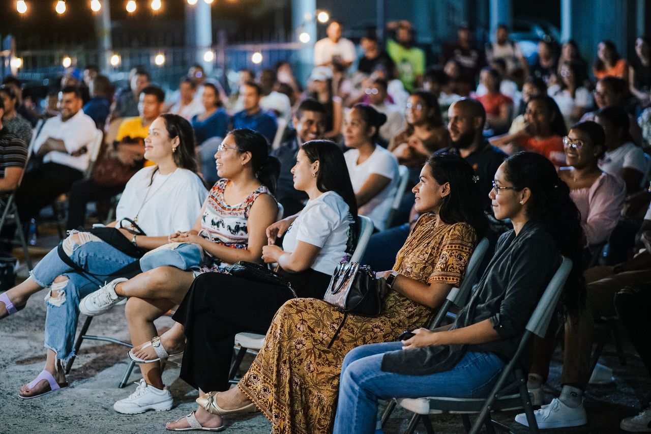A diverse group of adults attentively watching an outdoor event during the evening with ambient lighting.