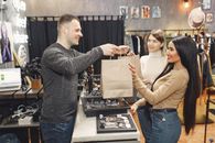 Side view of cheerful male shop assistant standing at counter desk and giving to positive female customers paper bags with purchases in trendy shop